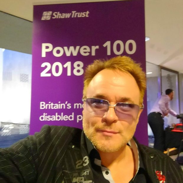 Mik poses in front of a purple backdrop, with the Shaw Trust Power 100 2018 on it