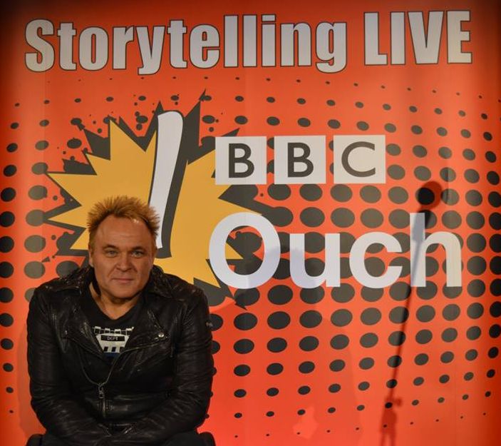 Mik smiling in a leather jacket, with a backdrop saying BBC Ouch Storytelling Live behind him.