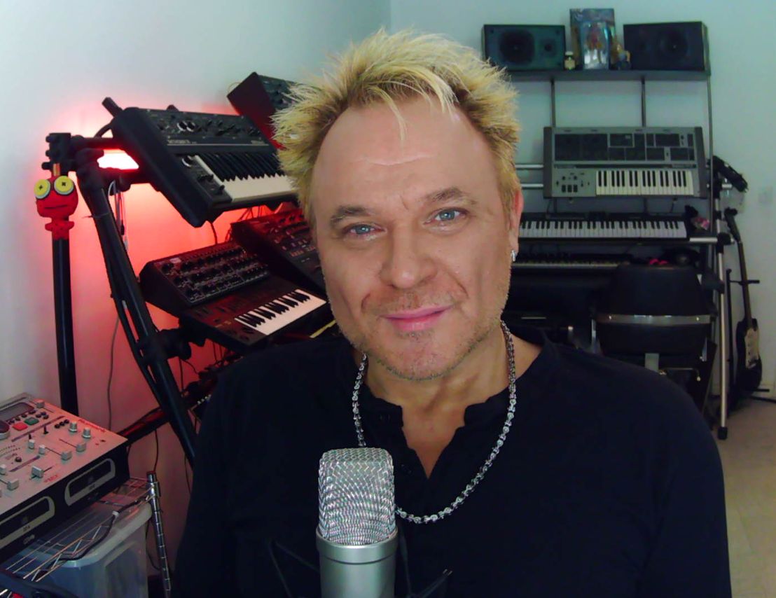 Mik in his studio, surrounded by musical equipment, with his mic in front of him. His lockdown zoom studio.